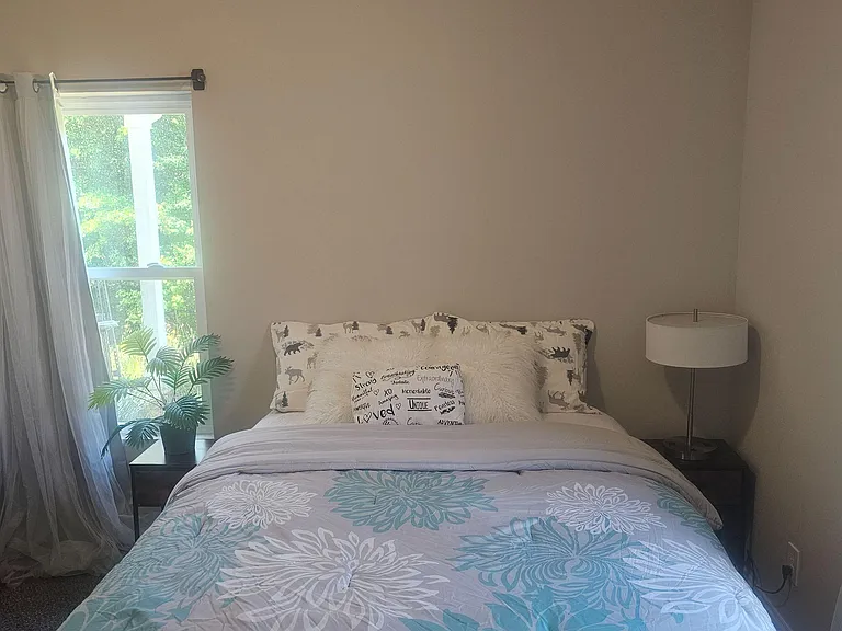 Furnished bedroom with a full-size bed, nightstand lighting, and large windows with curtains for natural light and privacy.