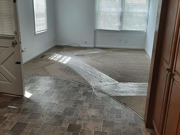 View from edge of kitchen into living room; front door is on the left in photo. Front of house and front yard is on south side of house, looking out the double windows. (Plastic runners are protecting carpet.)