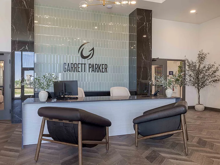The staffed front desk in the Garrett Parker leasing office welcomes prospective and current residents beneath a tiled accent wall displaying the community's logo.