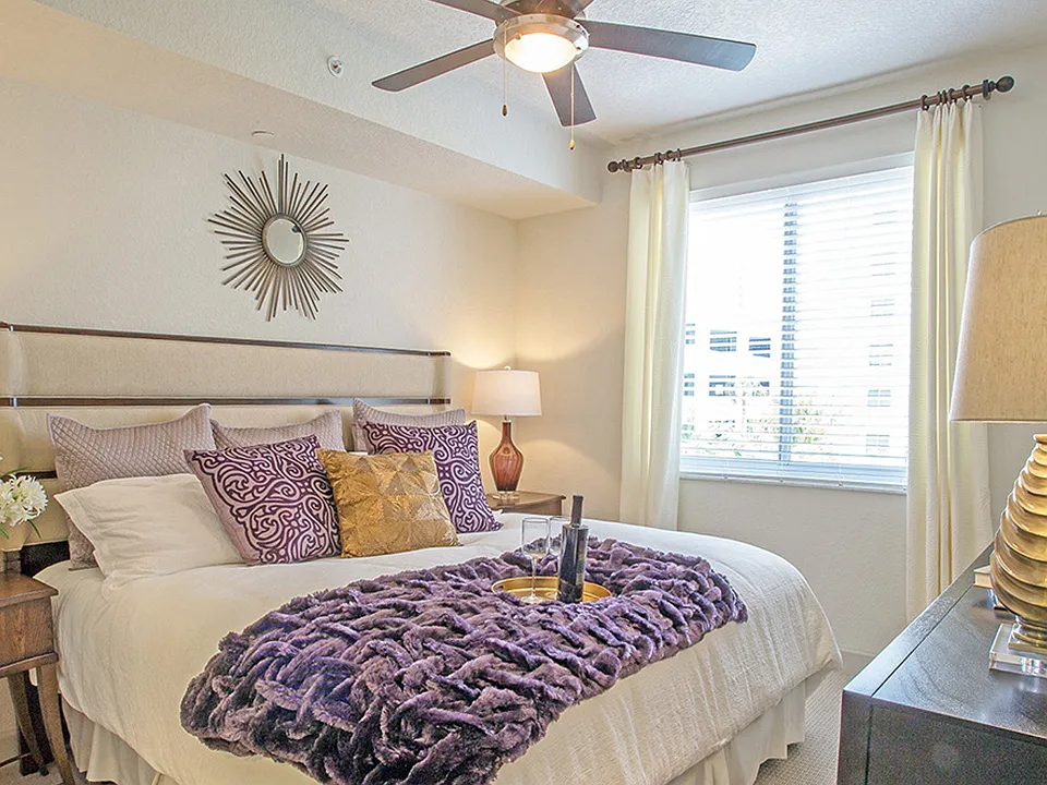 Luxury apartment bedroom with upholstered bed, ceiling fan, bedside lamps, and large window