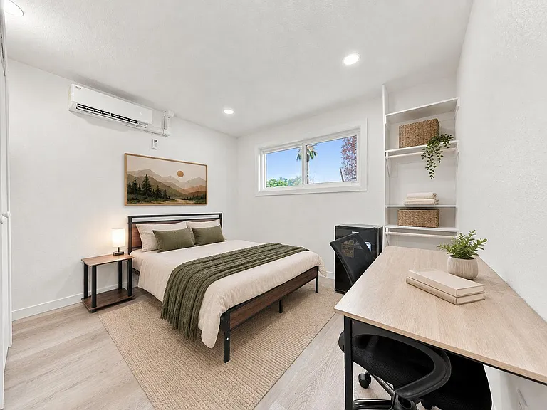Well-appointed bedroom with a full bed, dedicated desk workspace, open shelving, and a large window plus mini-split AC.