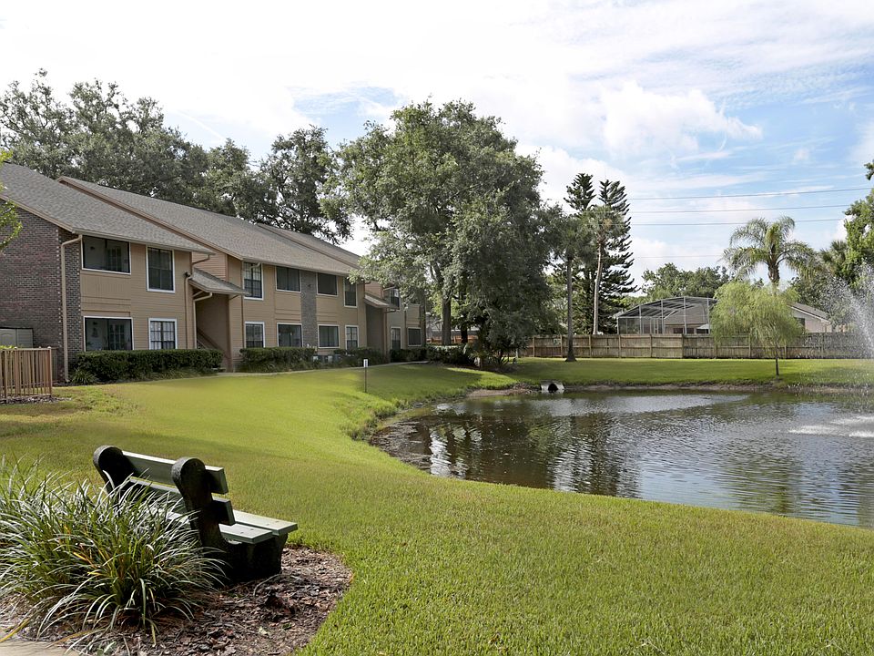 Park bench beside a grassy pond with a fountain, overlooking two-story brick-and-siding apartment buildings at The Park at Countryside.