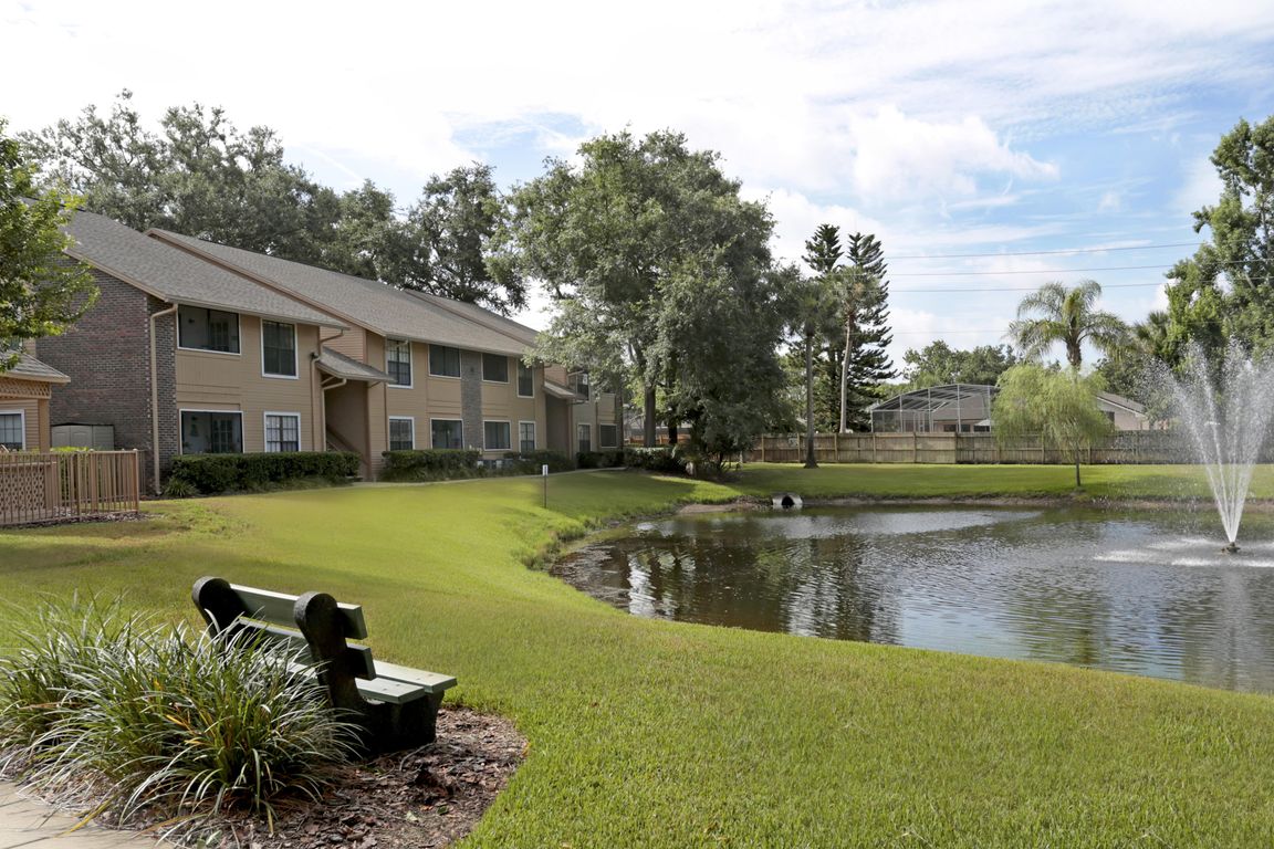 Park bench beside a grassy pond with a fountain, overlooking two-story brick-and-siding apartment buildings at The Park at Countryside.
