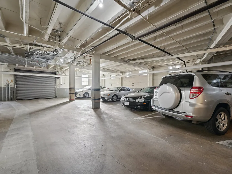 Underground parking garage with cars, marked spaces, lighting, and visible piping; closed shutter door