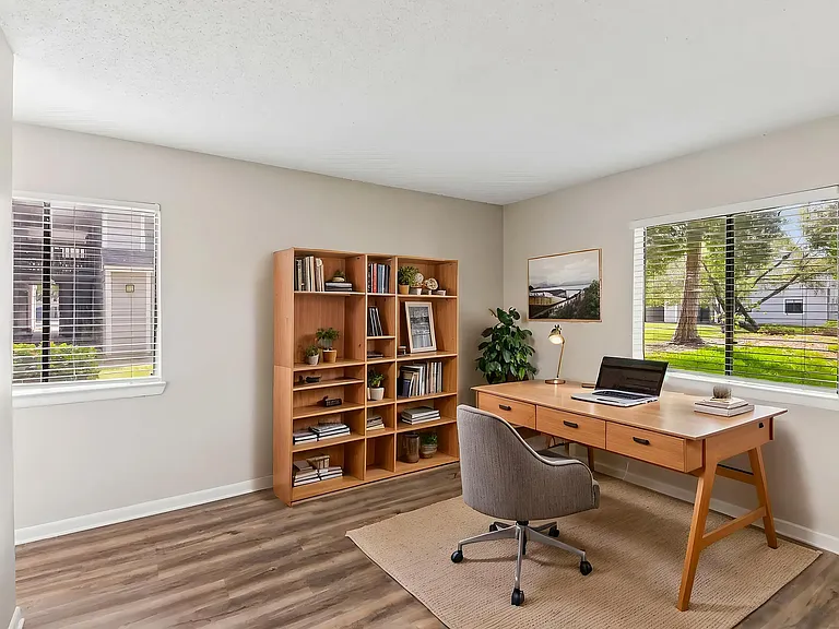 Bright, modern home office with large windows, wooden desk, and organized bookshelf, offering a serene view of lush greenery.