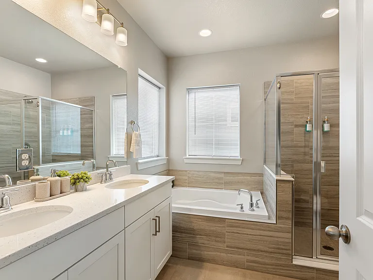 Bright modern bathroom with double vanity, white cabinets, and chrome fixtures. Features a large mirror, spacious bathtub with wood-look tile surround, and glass-enclosed shower. Natural light from two windows creates a clean, inviting space.