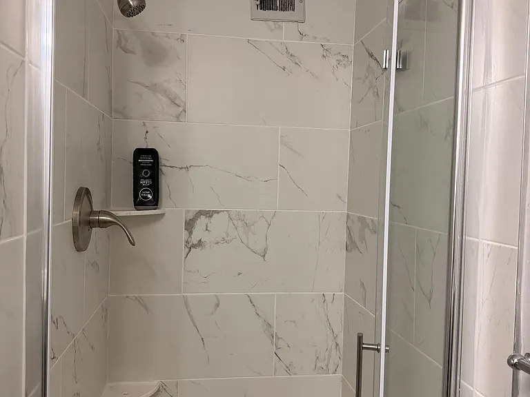 Walk-in shower with marble-look tile surround, glass door, built-in corner shelving, and modern shower fixtures.