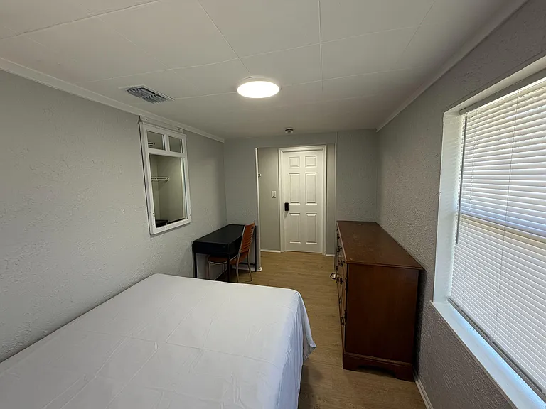 Private bedroom with a bed, dresser storage, and a dedicated desk area, plus a window with blinds for natural light.