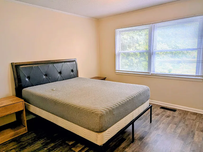 Spacious bedroom with a large window and blinds, plus room for a full/queen bed and nightstands on durable flooring.