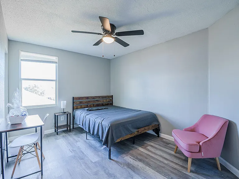 Private bedroom with a ceiling fan, large window, and wood-look flooring, plus a desk setup and accent chair for lounging.