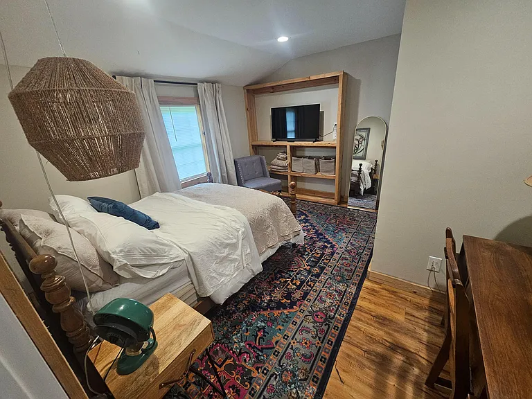 Furnished bedroom with a full bed, window with curtains, lounge chair, TV, and open shelving for storage.