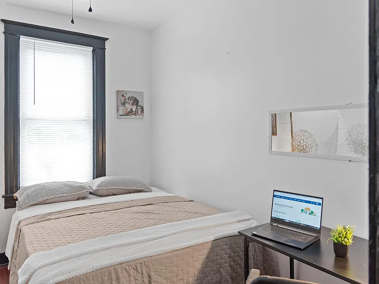 Furnished bedroom with a full-size bed, large window with blinds, and a compact desk area for work or study.