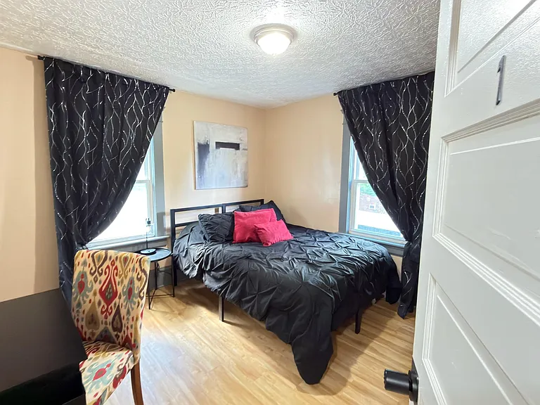 Furnished bedroom with a full bed, dual windows with blackout curtains, and hardwood floors for easy upkeep.