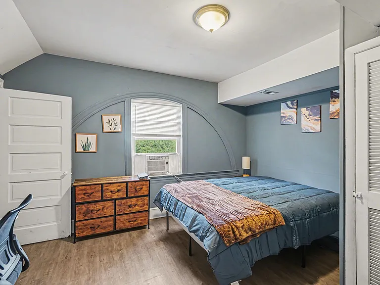 Furnished bedroom featuring a comfortable bed, dresser storage, and a window with blinds and in-room A/C unit.