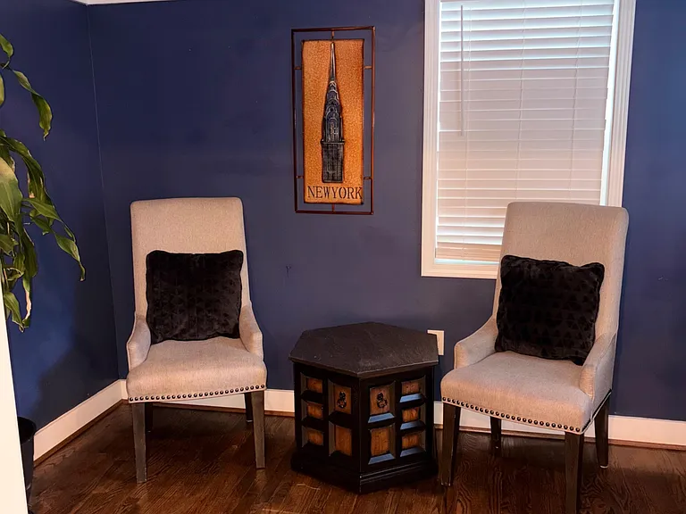 Dedicated sitting nook with two upholstered chairs, side table, and a window with blinds over hardwood flooring.
