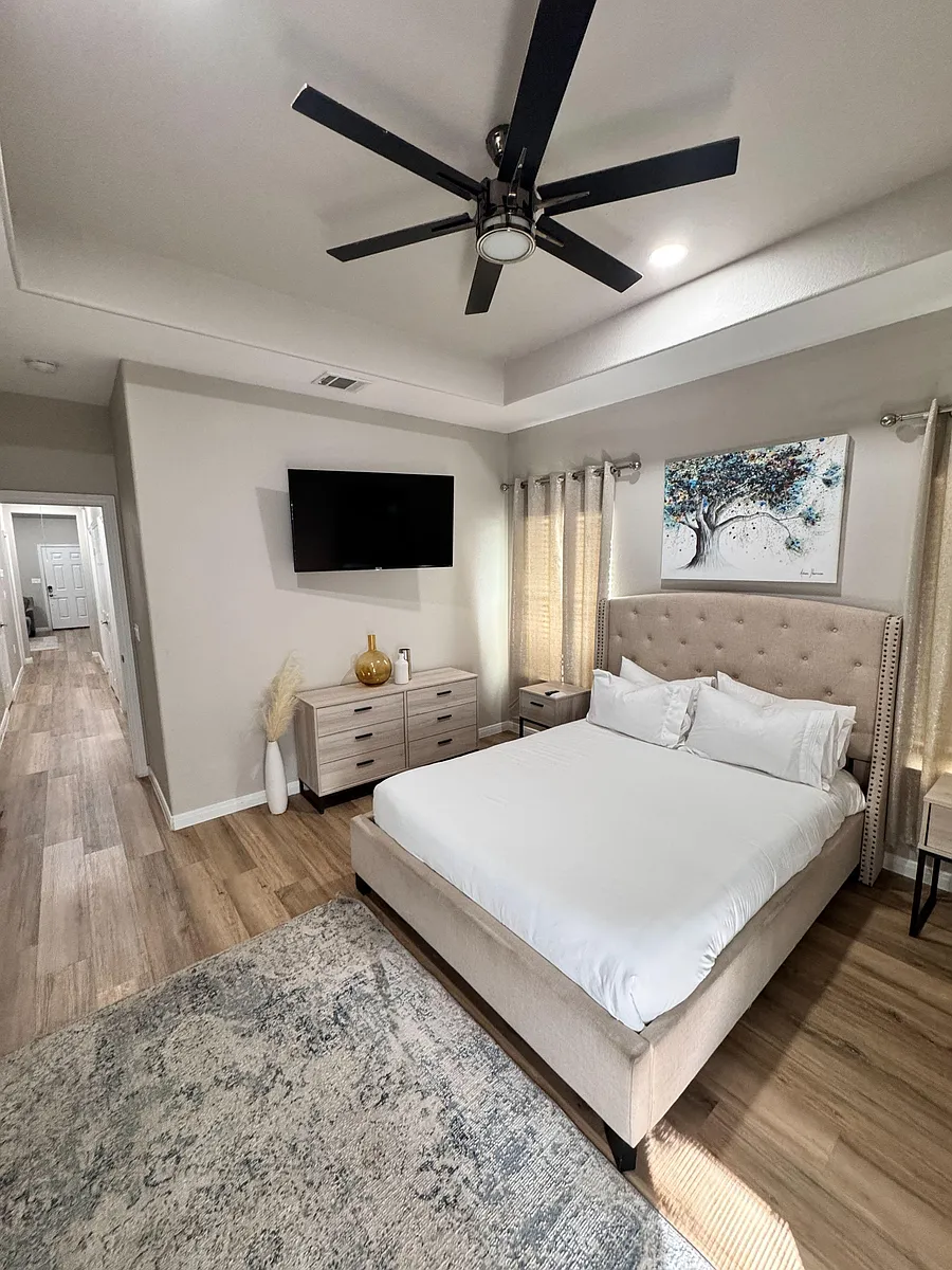 Spacious bedroom with a ceiling fan, mounted TV, dual nightstands, and a wide dresser, plus window treatments for privacy. - Calvin St