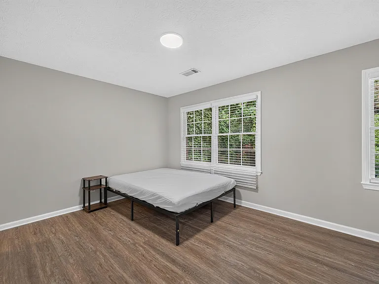 Spacious bedroom with wood-style flooring, neutral walls, and large windows with blinds overlooking greenery.