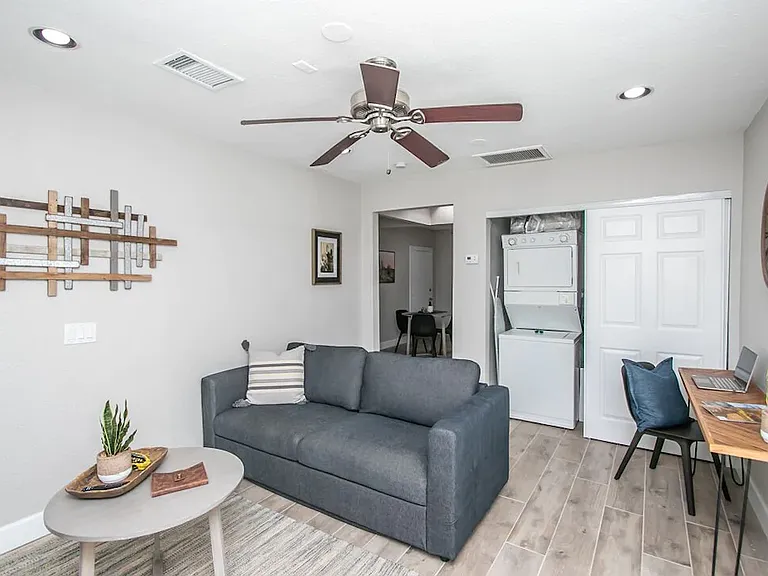 Detached casita living area at, Tempe rental home. Features dark grey pullout sofa, coffee table, wood-plank style flooring, a dedicated work desk with a laptop, and a convenient stacked washer/dryer unit. Cozy, functional space for a top Tempe, AZ stay.