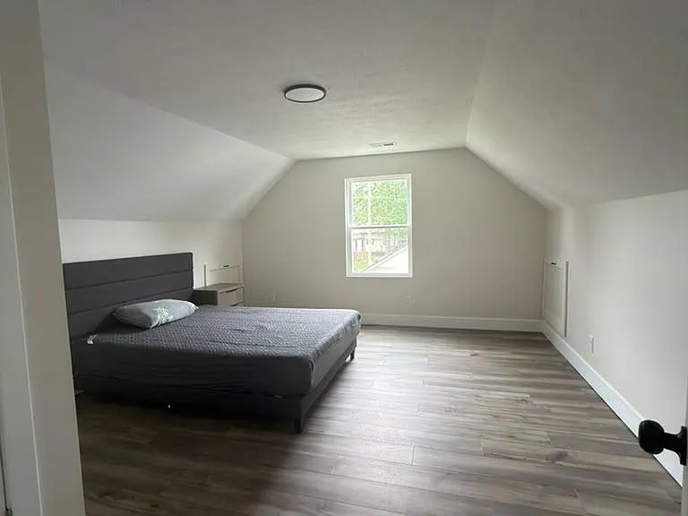 Spacious loft-style bedroom with sloped ceilings, wide-plank flooring, and a centered window for natural light.