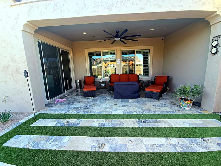 This covered backyard lounge features cushioned seating, ottomans, and a fire table at the center. Travertine tiles with grass accents, a ceiling fan, and playful decor create a breezy retreat perfect for outdoor relaxation and evening ambiance.