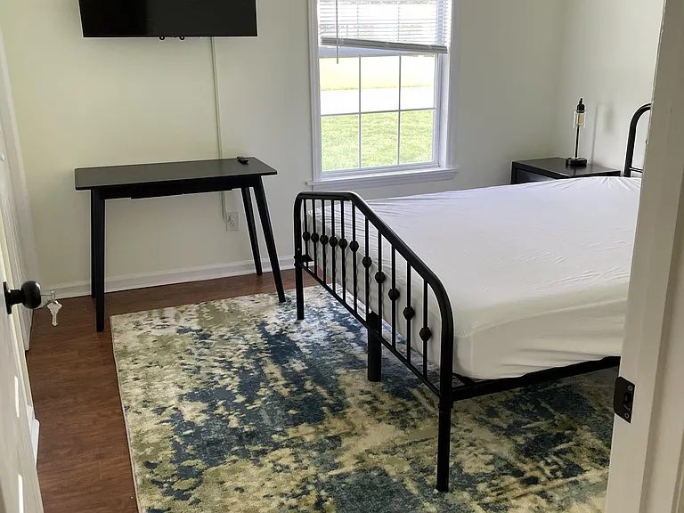 Furnished bedroom with a full bed, desk, and wall-mounted TV, plus a large window with blinds and area rug.