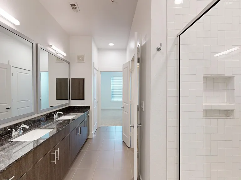 Apartment bathroom with glass-enclosed shower, tile flooring, and double sinks.