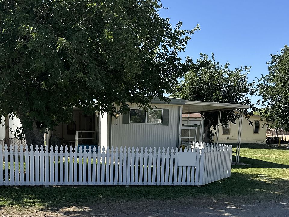 Front of mobile home with fenced yard including a large shade tree and plenty of parking area in the grass to the right.