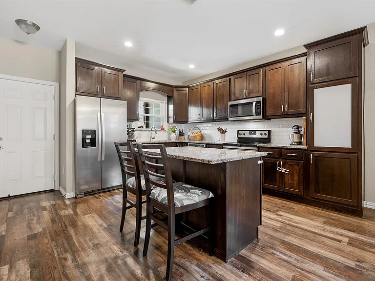 Kitchen featuring wood-finish flooring, dark wood cabinetry, a center island with a granite-style countertop, stainless steel appliances, and a white subway tile backsplash