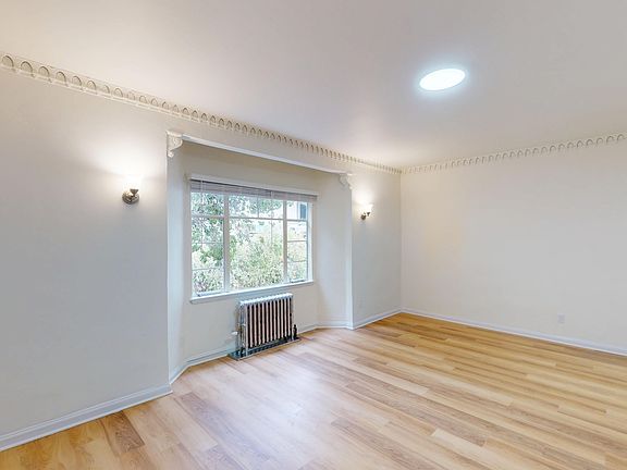 Bright, empty room featuring wooden flooring, a large window, and decorative crown molding, a radiator is visible beneath the window