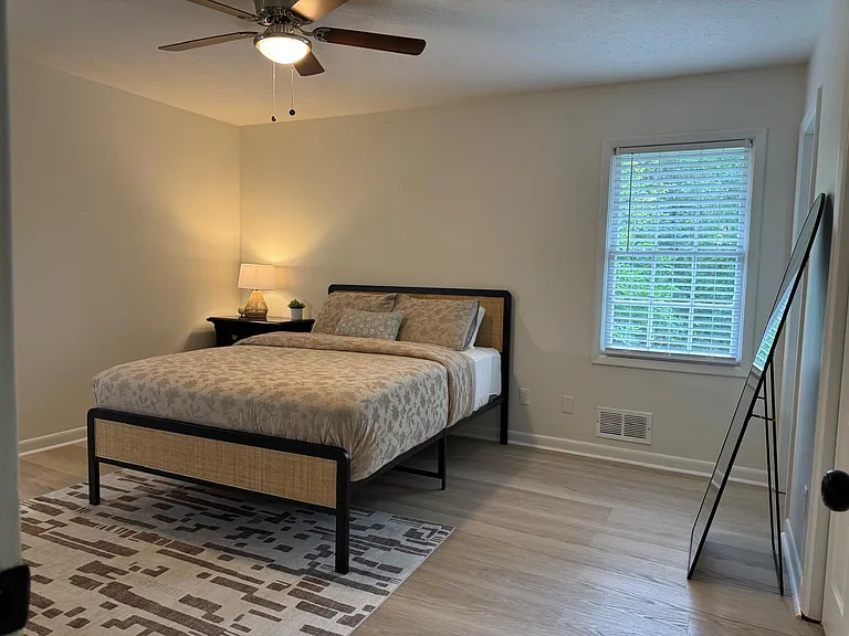 Furnished bedroom with a queen bed, nightstand, ceiling fan, and window with blinds for privacy and natural light.