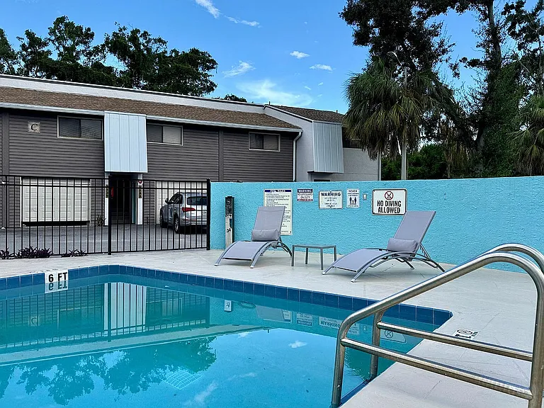 Inviting pool area with relaxing lounge chairs in a serene apartment complex setting, enhanced by lush greenery.
