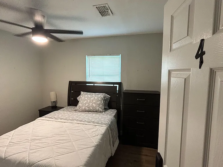 Furnished bedroom with a bed, nightstand, and dresser, plus a ceiling fan, mini fridge and all bedding.