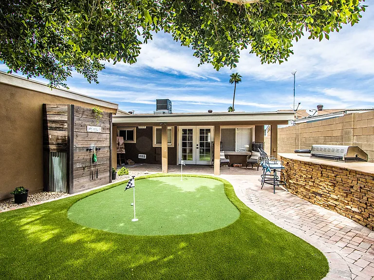 Scenic backyard view at Casa Del Sol, Tempe rental home. Features a custom putting green with golf flags, a stone BBQ island with bar seating, brick pavers, a covered patio, and putter storage. Perfect outdoor entertainment setup for a Tempe, AZ stay.