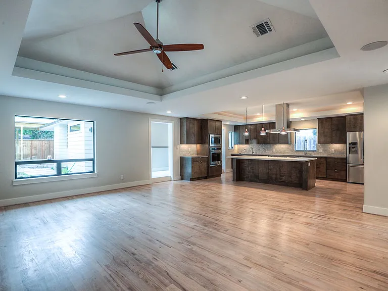 Open living/kitchen view of vaulted tray ceiling with ceiling fan, large picture window, and oversized island kitchen with pendant lighting