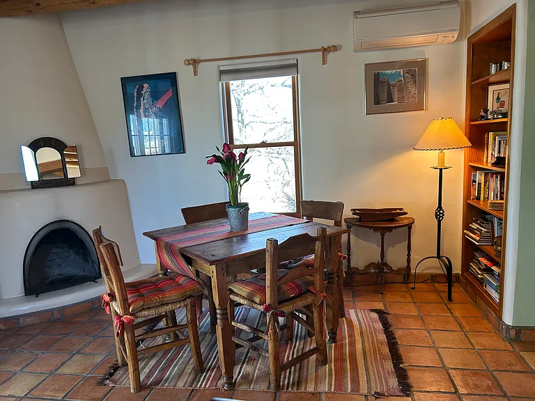Dining area of the Nambe Casita with kiva fireplace and small library. Window looks out towards old apricot tree and acequia.