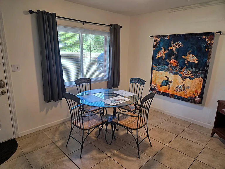 Dining area with a round glass-top table for four, tile flooring, and a large window with blinds and curtains.