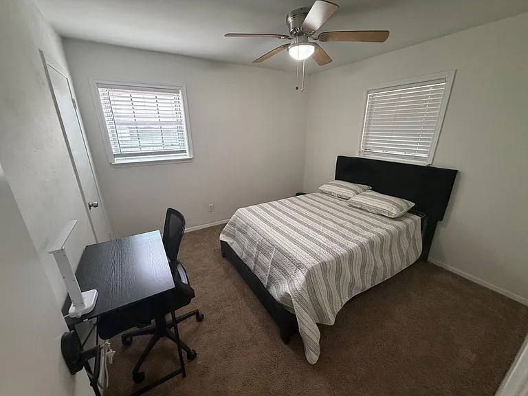 Furnished bedroom with a full-size bed, work desk, ceiling fan, and two windows with blinds for natural light.