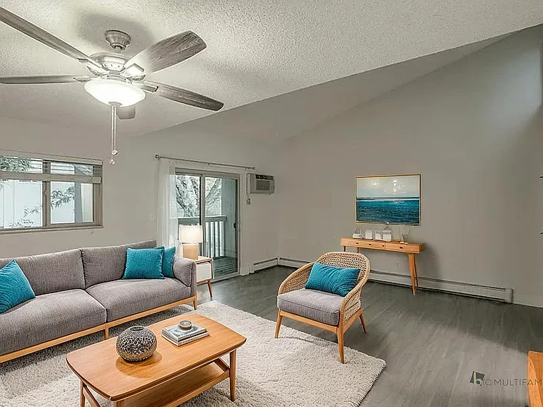 Inviting living room with modern gray sofa, vibrant teal cushions, and stylish wicker chair, enhanced by chic decor and natural light from large windows.