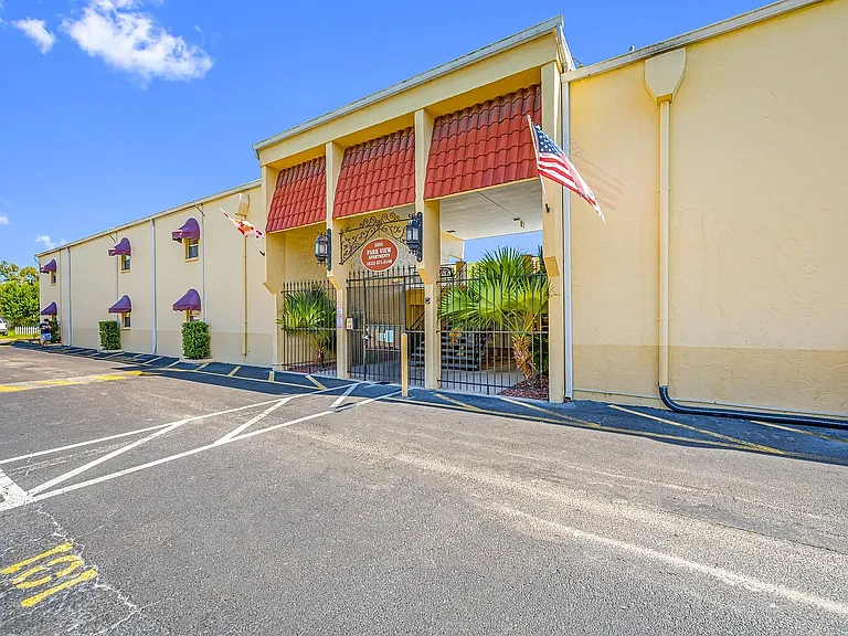 Charming apartment complex with striking red tile roof, welcoming entrance, and lush palm landscaping under a bright blue sky.