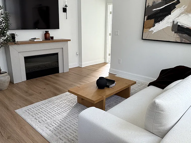 The apartment offers a warm, modern organic feel with an electric fireplace, high ceilings, and large loft-style windows that create a bright and airy living space.