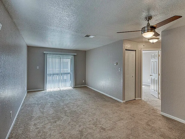 Bright and spacious living room with plush carpet, modern ceiling fan, and expansive windows with vertical blinds.