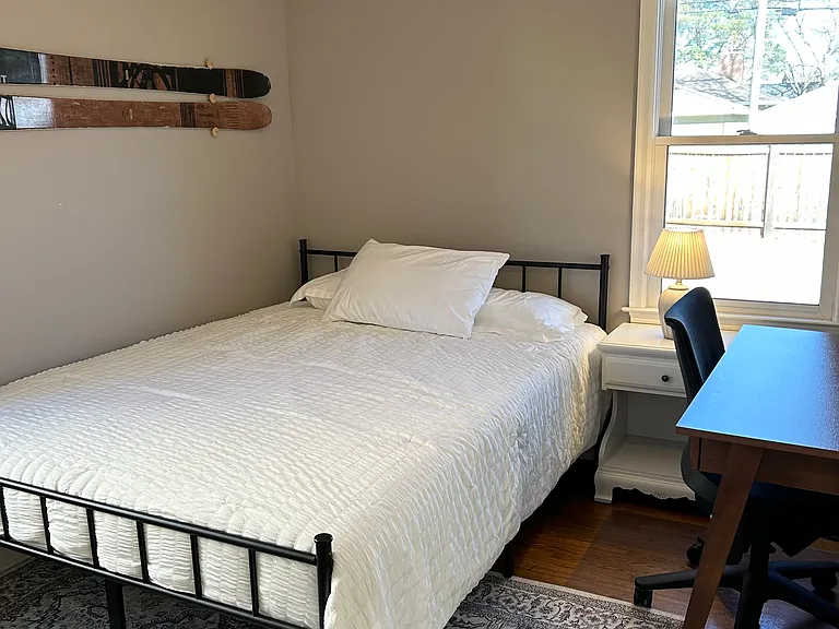 Main floor bedroom 1 features a queen size bed, desk and chair, nightstand, lamp, closet with a laundry basket and hangers.