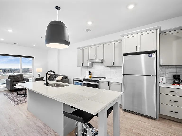 Modern open-concept kitchen and living area with large windows, stylish pendant light, and sleek stainless steel appliances. Modern open-concept kitchen and living area with large windows, stylish pendant light, and sleek stainless steel appliances.