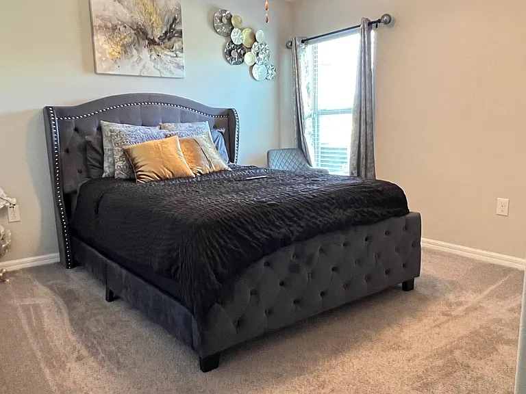 Spacious carpeted bedroom with a queen bed, upholstered headboard, and a large window with curtains for natural light.