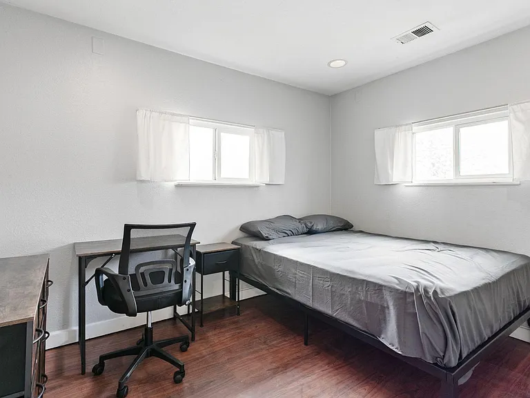 Furnished bedroom with a bed, nightstand, and dedicated desk area, plus two windows for natural light.