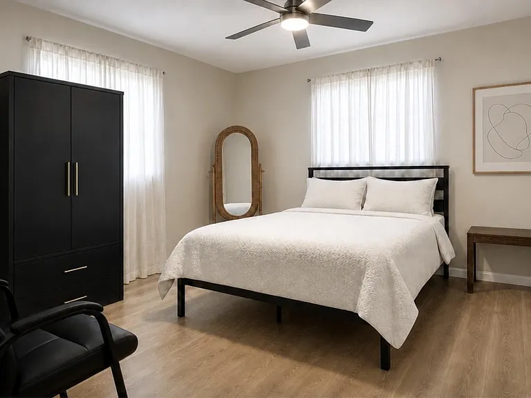 Spacious bedroom with a queen bed, large wardrobe, ceiling fan, and two windows with sheer curtains for soft natural light.