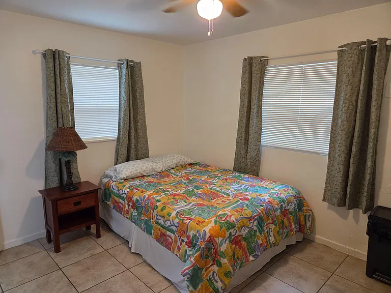 Furnished bedroom featuring a queen-size bed, nightstand, and two windows with blinds and curtains for privacy. paddle fan