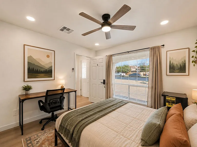 Well-appointed bedroom with a work desk, ceiling fan, and large window with blinds and curtains for light control.