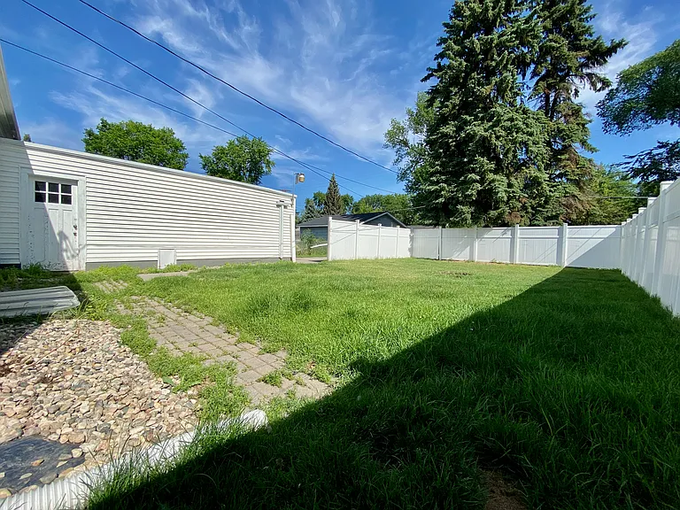Spacious backyard with lush green lawn, bordered by a white picket fence, and a charming white exterior building under a clear blue sky. Spacious backyard with lush green lawn, bordered by a white picket fence, and a charming white exterior building under