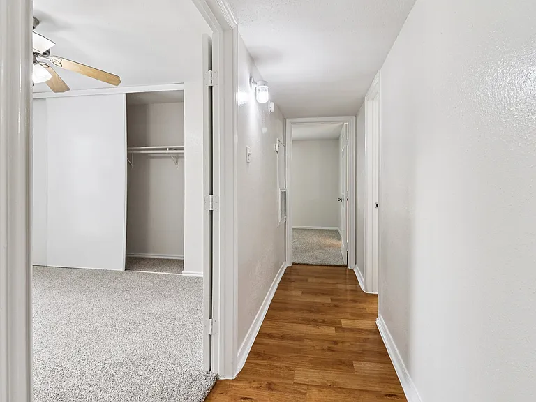 Bright hallway with fresh white walls, wood-style floor, and open closet space leading to a cozy carpeted bedroom with a ceiling fan. Bright hallway with fresh white walls, wood-style floor, and open closet space leading to a cozy carpeted bedroom with a 